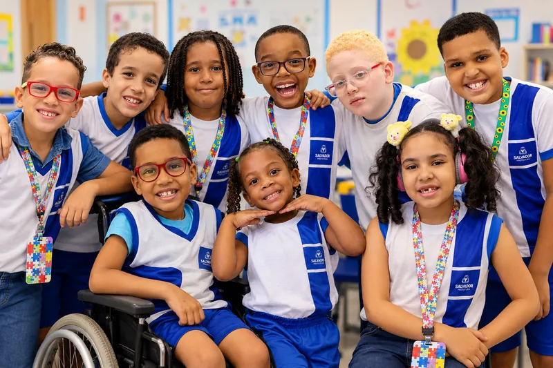 Grupo de estudantes sorrindo na sala de aula, com o uniforme da rede municipal de Salvador, um deles em cadeira de rodas