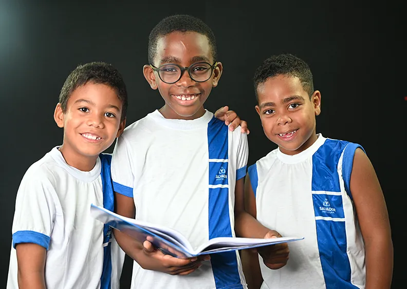 Três estudantes de uniforme sorrindo e segurando juntos um livro aberto